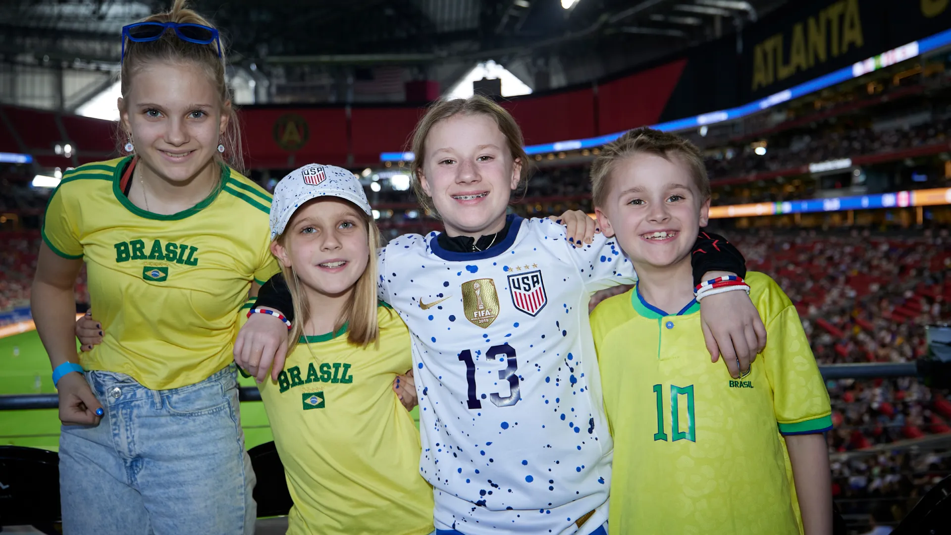Four kids smiling at a stadium wearing Brazil and USA soccer jerseys, with Atlanta sign in background.