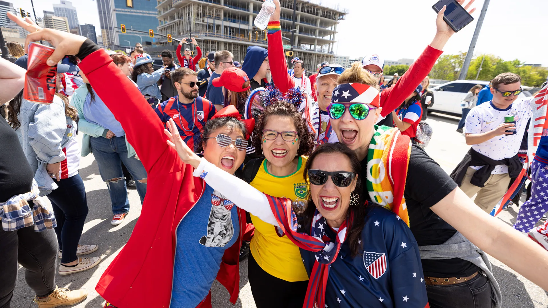 Group of excited soccer fans in colorful jerseys and sunglasses celebrating outdoors on a sunny day.