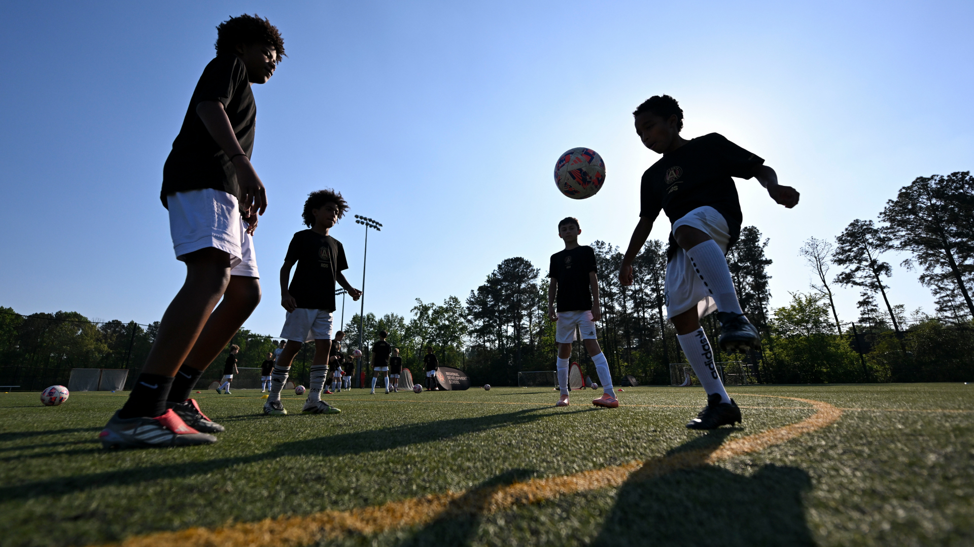 Young children in black shirts and white shorts practicing soccer on a field with trees and clear sky.