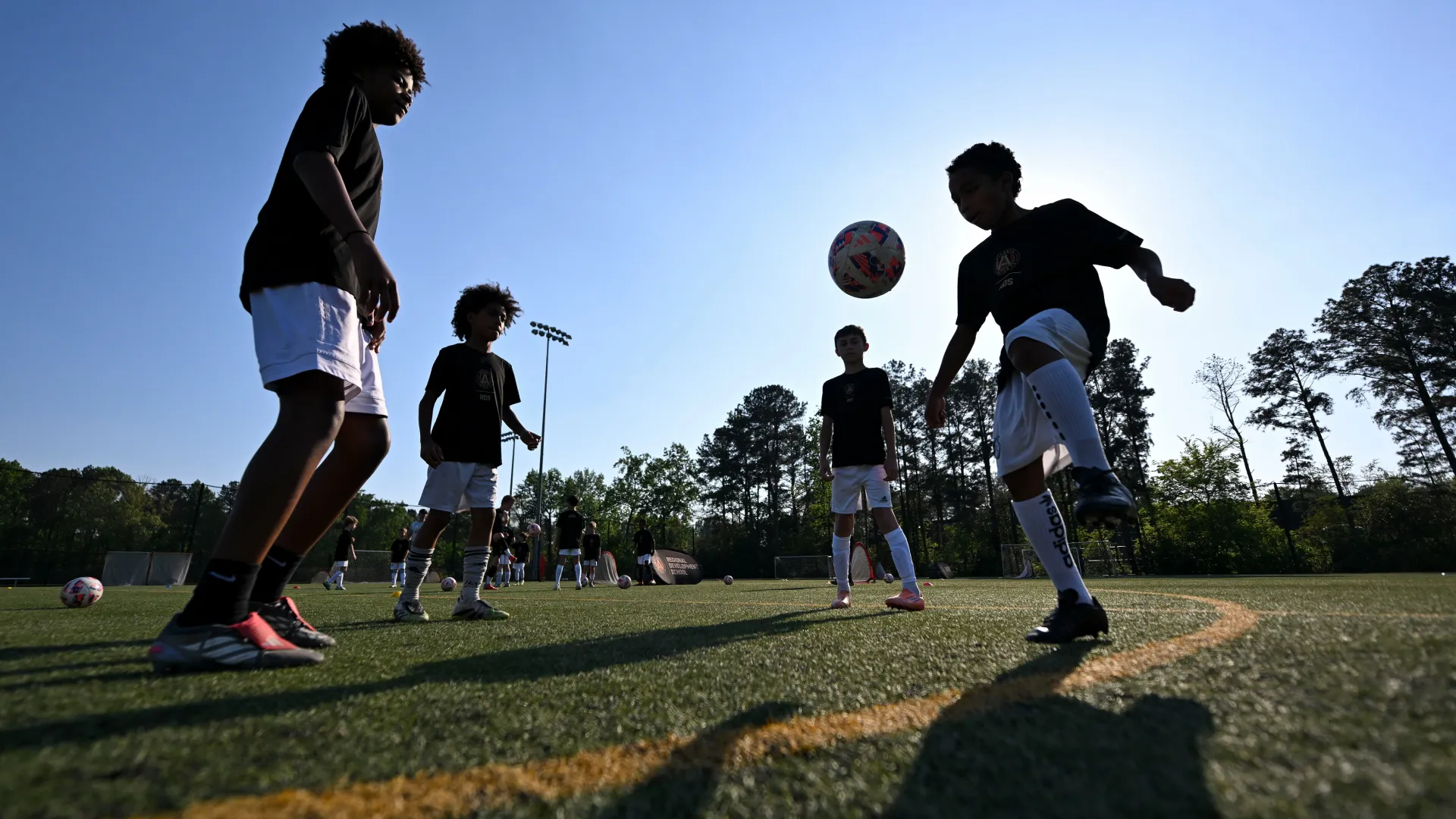 Young children in black shirts and white shorts practicing soccer on a field with trees and clear sky.