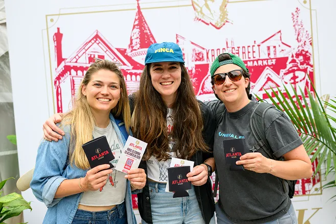 Three women smiling together at an outdoor Atlanta street festival, holding event wristbands and koozies, with a red-and-white illustrated mural in the background.