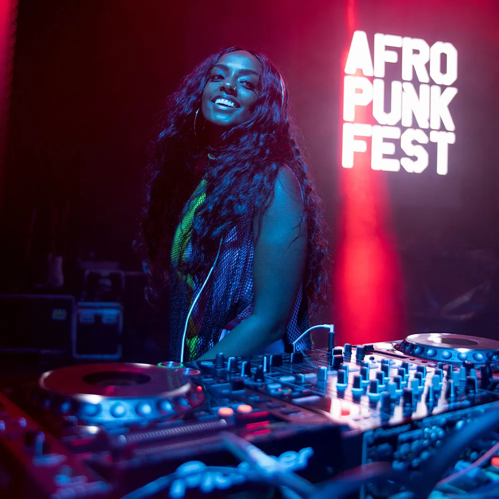 Osho smiles behind a DJ mixer in front of a neon Afropunk Fest sign.