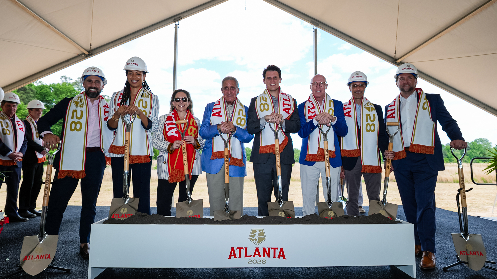 Officials in hard hats and Atlanta 2028 scarves hold ceremonial shovels at a groundbreaking ceremony.