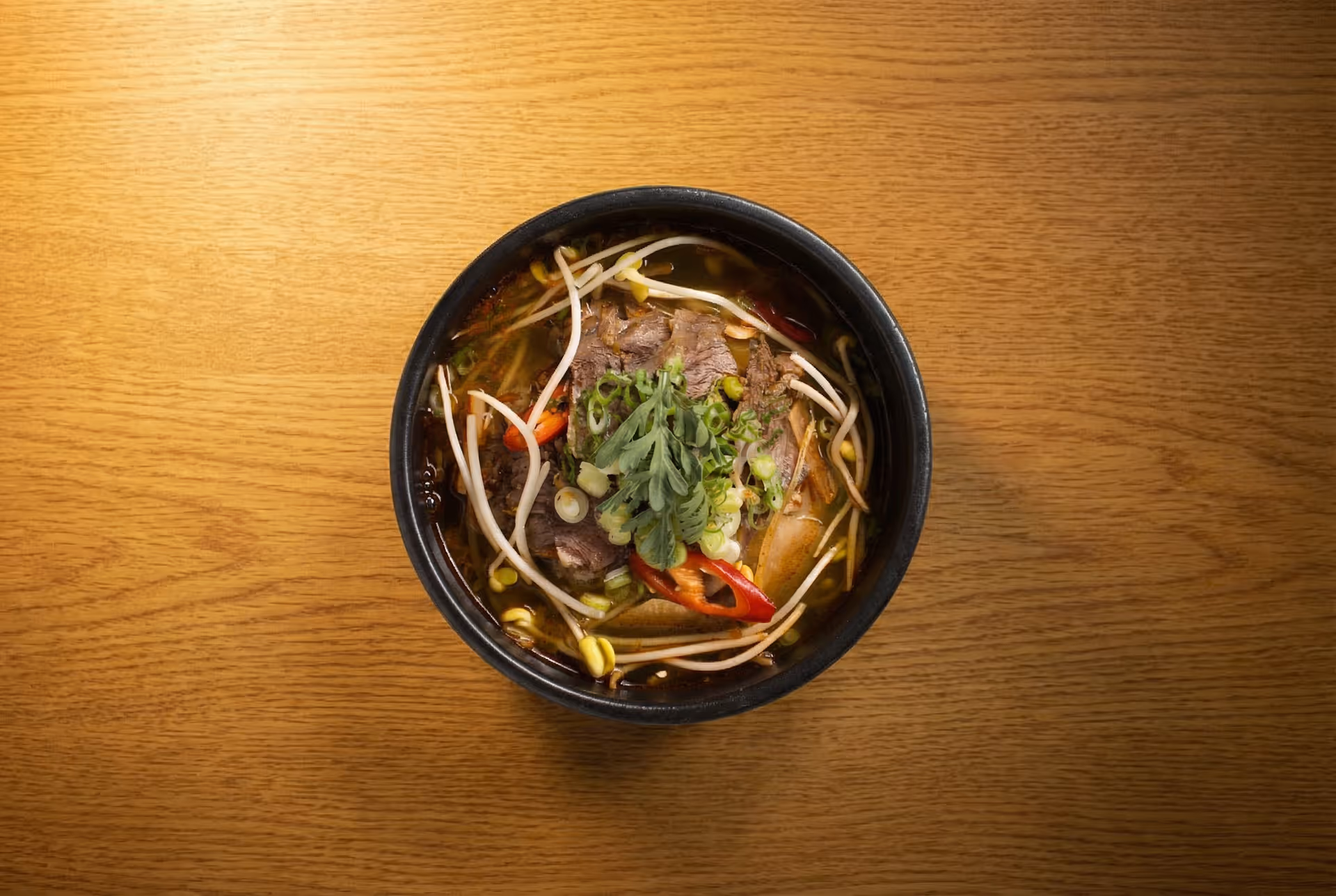 Top-down view of a bowl of beef noodle soup with bean sprouts, chilies, scallions, and fresh herbs on a wooden table.