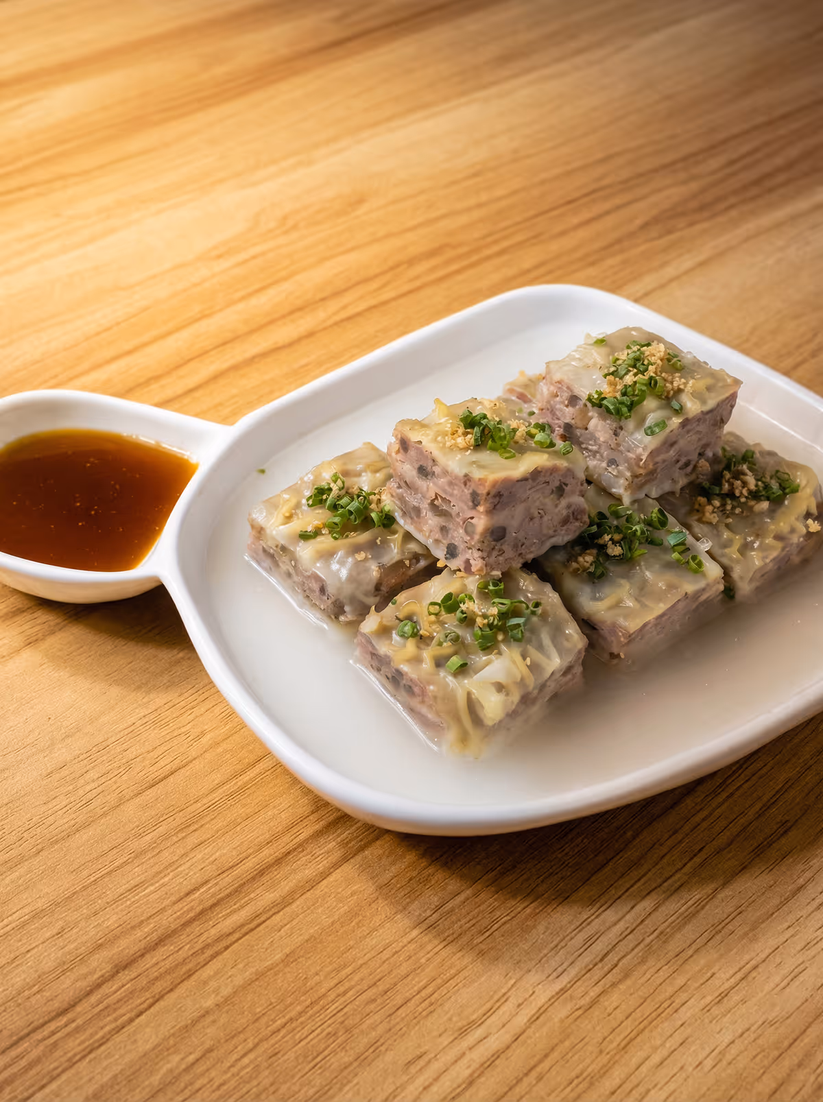 White dish with six square pieces of steamed meat topped with chopped green onions and served with a brown dipping sauce in a small compartment.