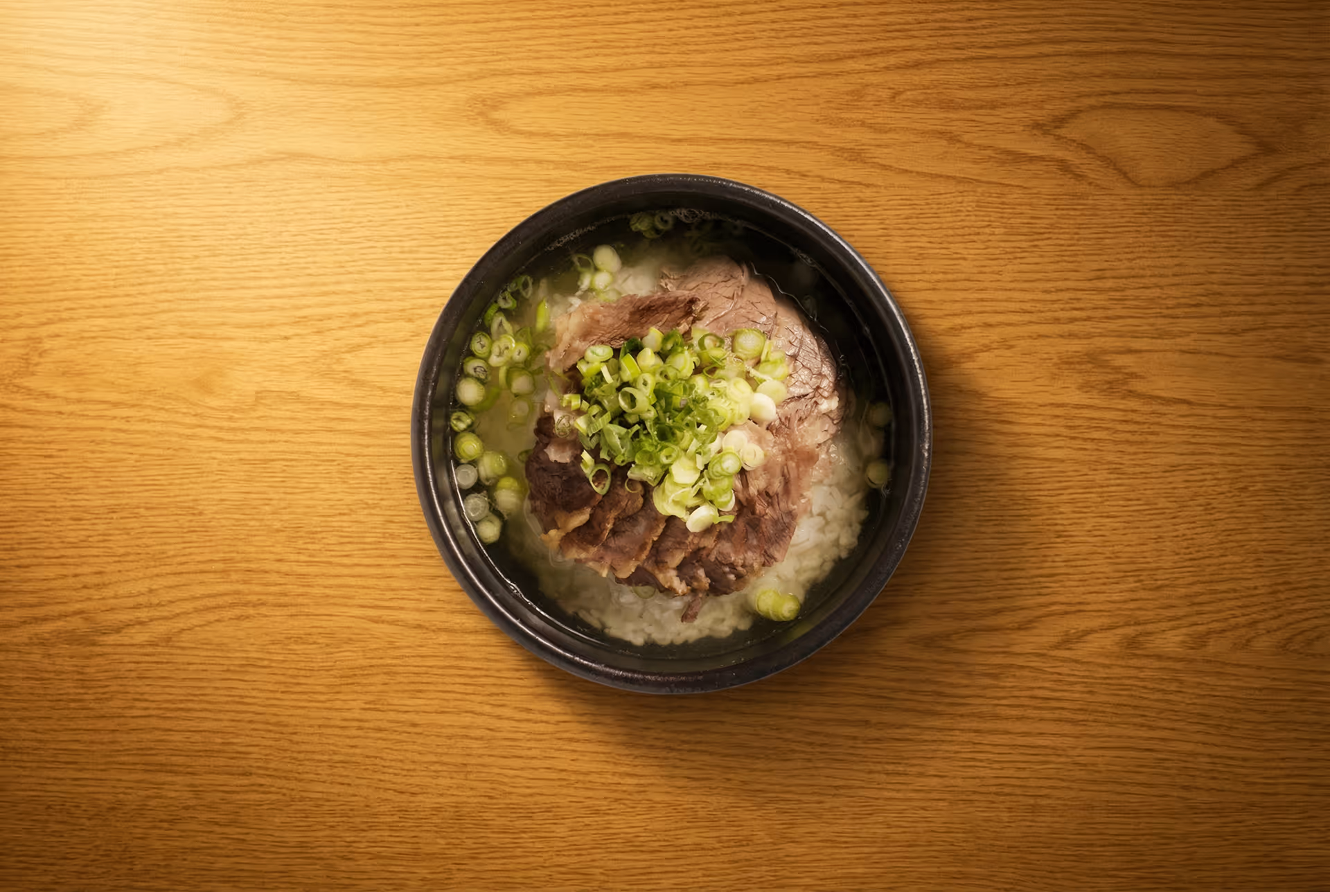Bowl of soup with slices of beef, rice, and chopped green onions on a wooden surface.