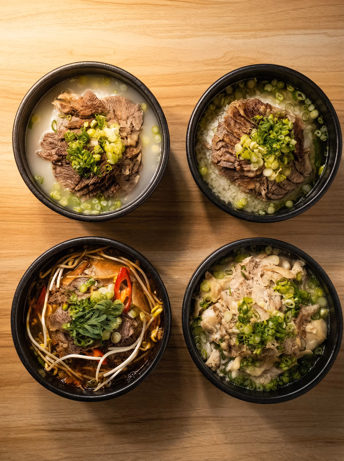Four bowls of Asian soup with sliced beef, topped with chopped green onions and herbs, arranged in a square on a wooden surface.