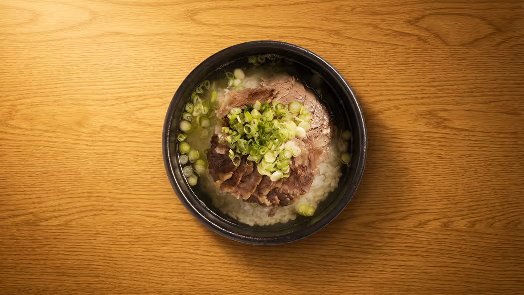 Black bowl on wooden table filled with soup containing sliced beef, chopped green onions, and rice.