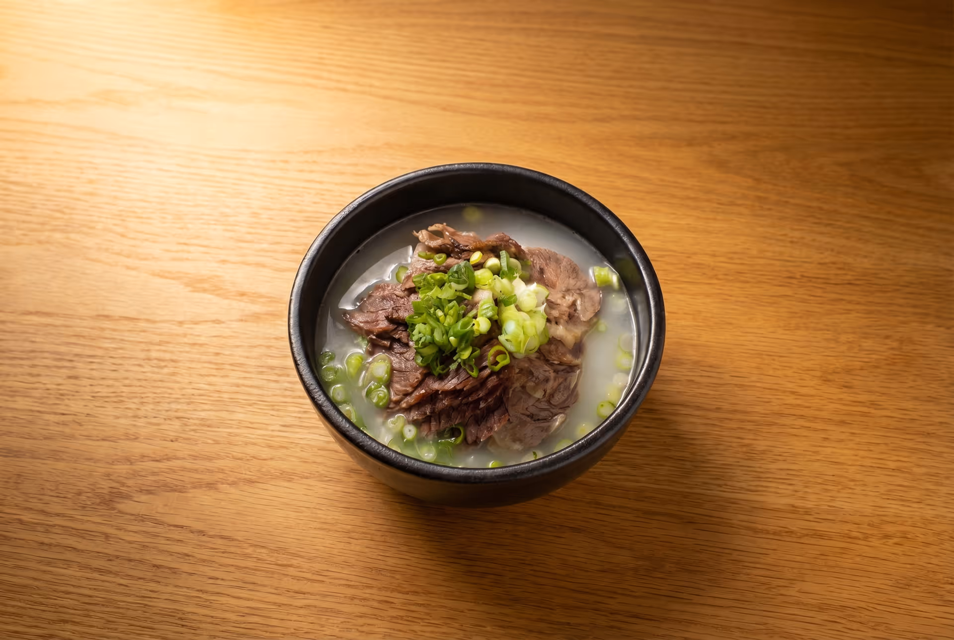 Black bowl filled with beef soup garnished with chopped green onions on a wooden table.
