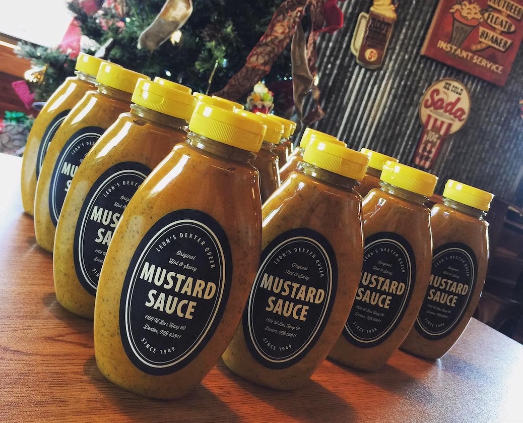 Rows of mustard sauce bottles with yellow caps on a wooden table in a rustic setting.