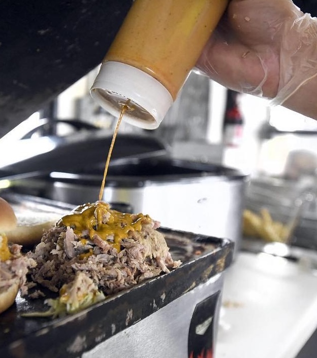 Person wearing a glove pouring mustard sauce on a sandwich pile of shredded meat on a black tray in a kitchen.