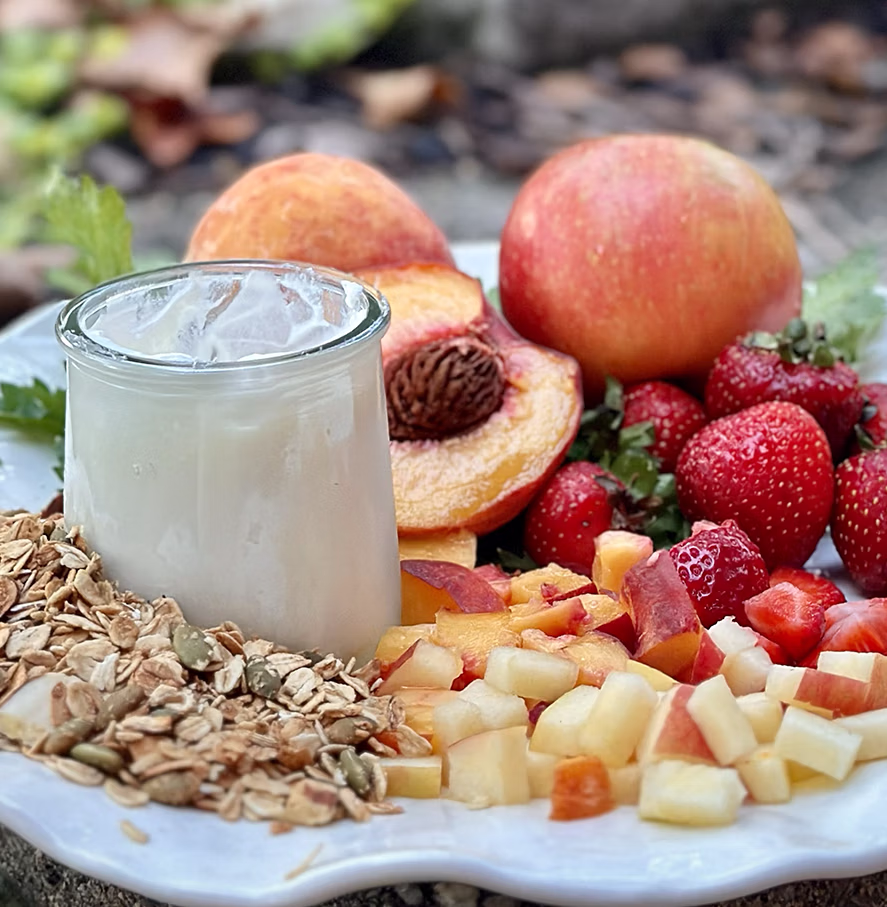 Plate with granola, a glass of yogurt, whole and sliced peaches, chopped apples, and strawberries.