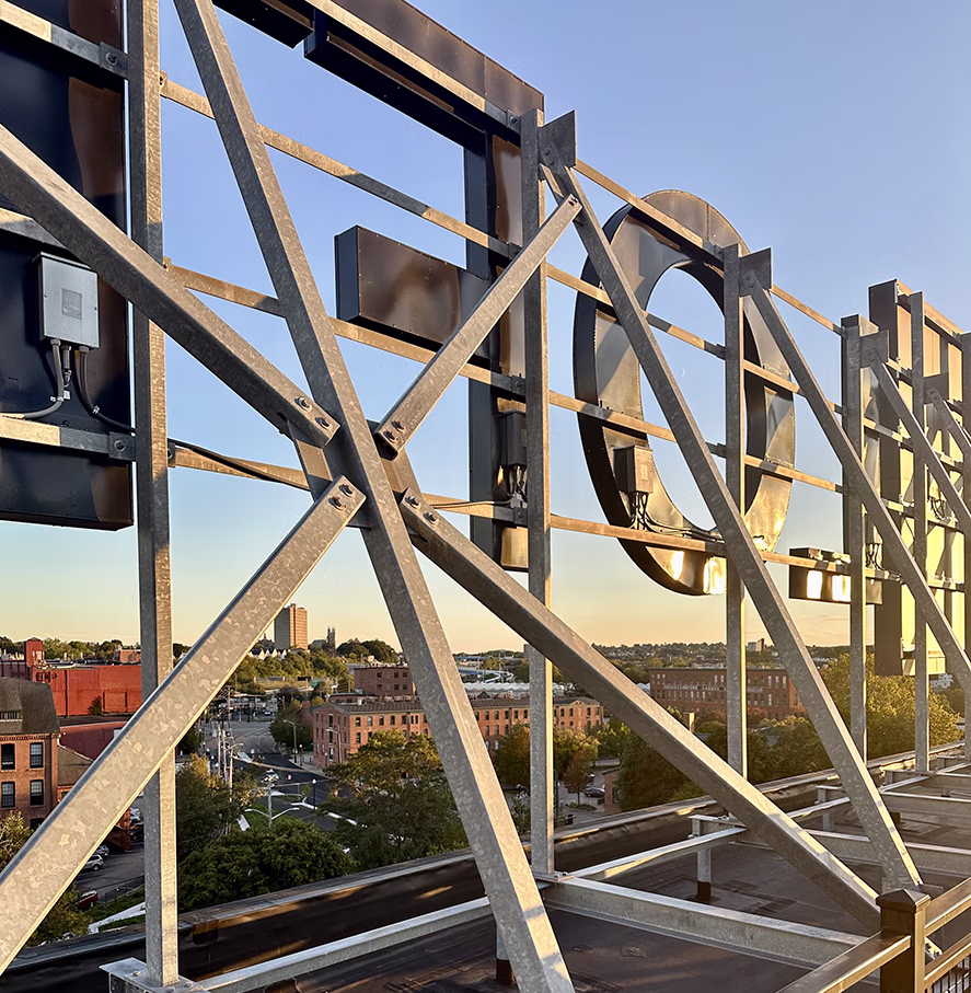 Back view of a large metal rooftop sign structure overlooking a cityscape with buildings and trees under a clear sky at sunset.