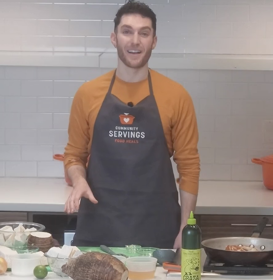 Jason wearing a Community Servings apron stands in a kitchen behind a counter with various cooking ingredients and utensils.