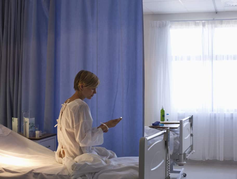 A female patient with short blond hair sits on a hospital bed and looks down at her cell phone.