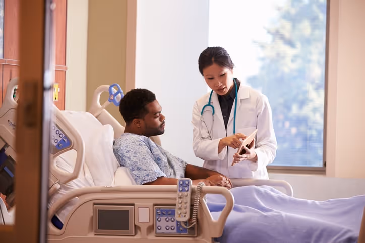 An African-American male patient sits in a hospital bed as an Asian doctor explains what is on her iPad screen.