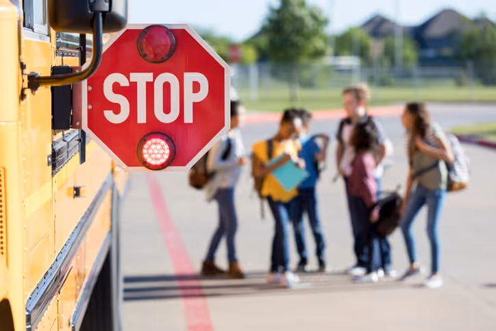 A closeup shot of a stop sign on a school bus shows the flashing red lights as children leave the bus in the background.