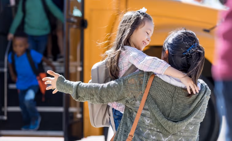 A mother squats down with her arms open to embrace her daughter as she gets off the school bus.