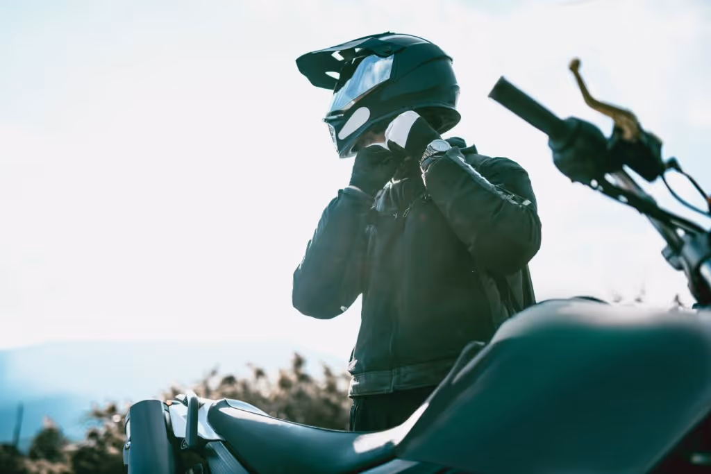 Man with black jacket and gloves adjusting strap of black motorcycle helmet in front of black motorcycle