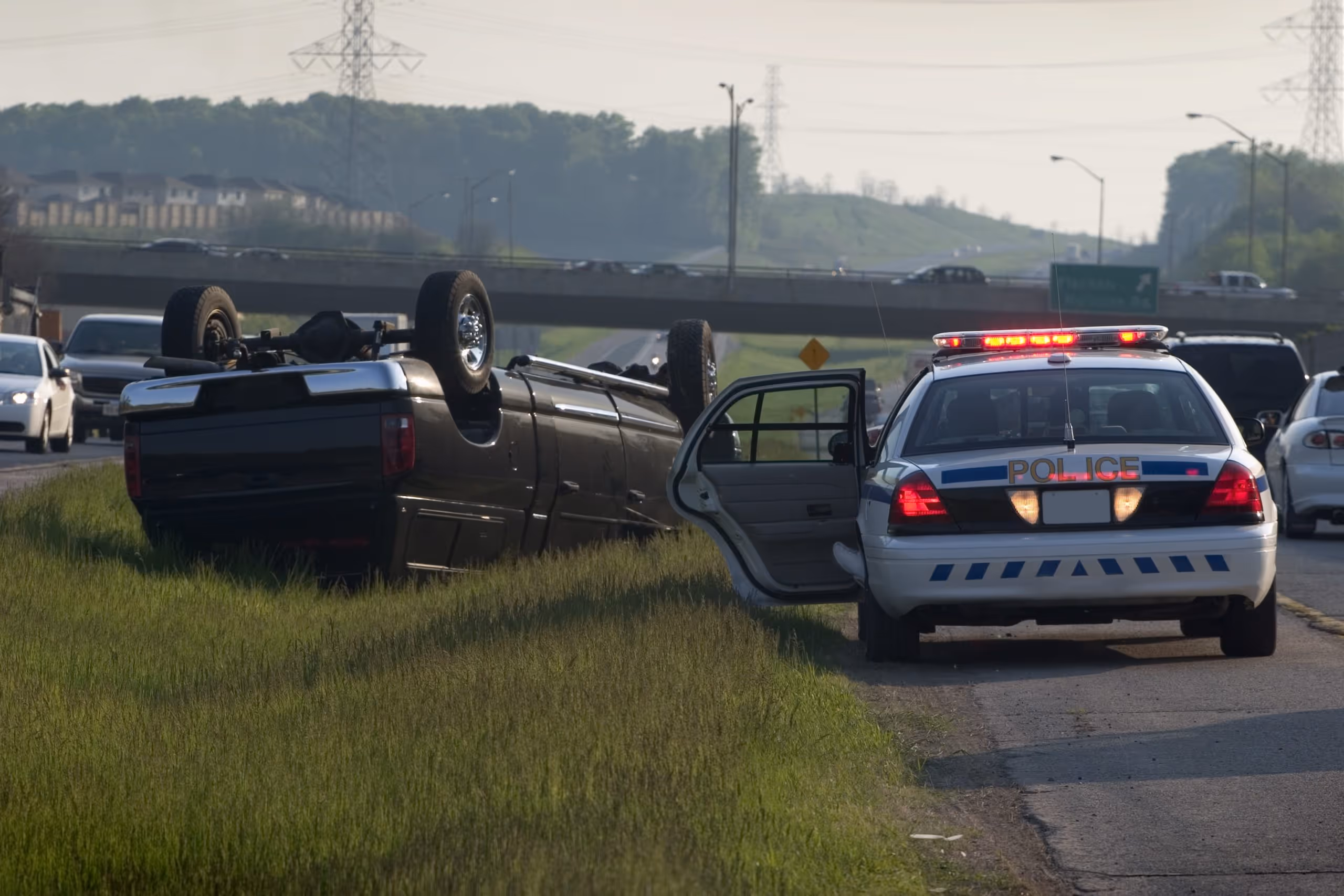 Black suv upside down in the grass after a rollover accident, attended to by local police|Freeway sign showing possible truck rollover accident dangers|A winter scene on a of a car that has overturned after a rollover accident due to treacherous driving conditions. The shot was taken as the sun was rising to reveal the ice and snow from the previous night was starting to thaw. Very Overcast sky. No visible manufacturer logos.