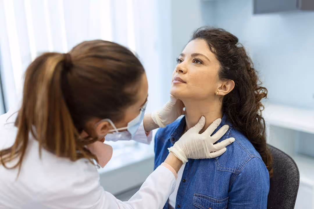 A woman with dark curly hair wearing a light blue blouse lifts her head so a doctor can evaluate her medical condition.|Nurse in green scrubs holding a medical form with blurred patient in background