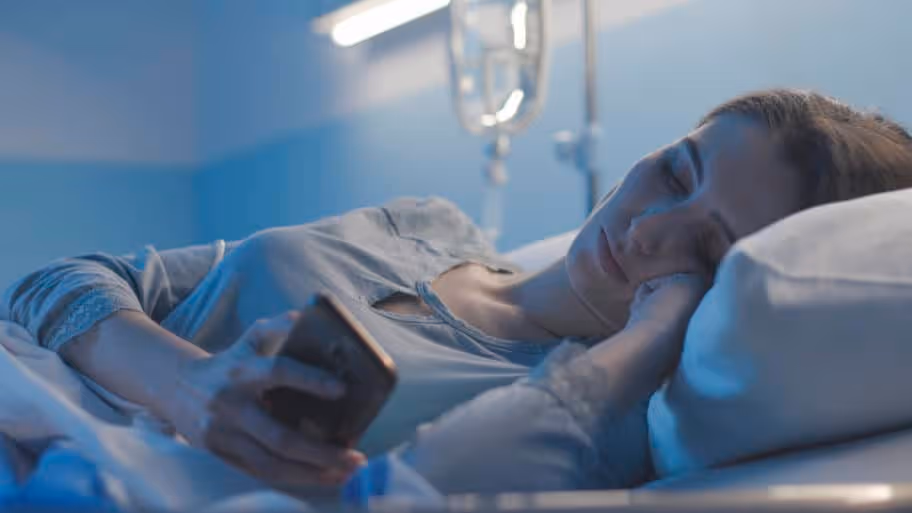A sleepless female patient searches her phone while laying on a hospital bed in bluish light.