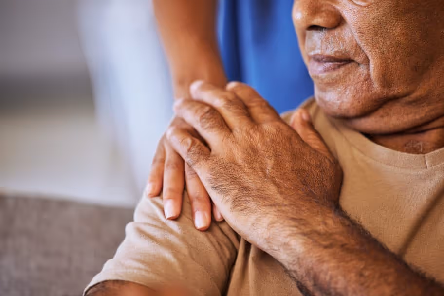 A patient and his caregiver grasp hands over his shoulder.