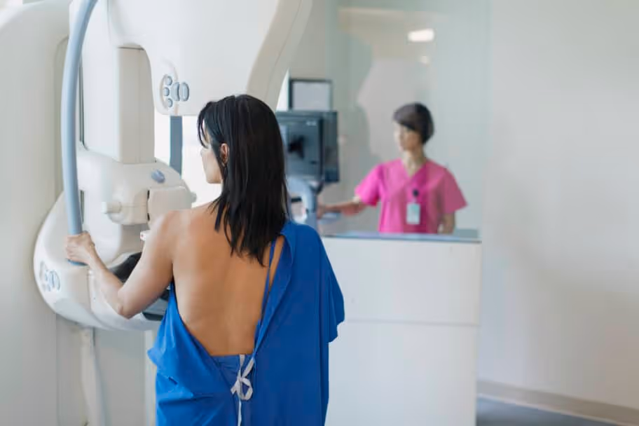 A woman with dark hair undergoes a mammogram under the direction of a nurse in pink scrubs.