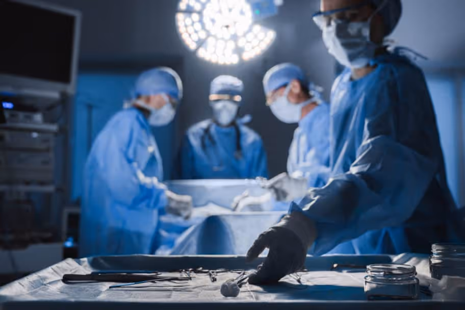 A bluish image of surgeons operating in the background while one doctor in scrubs, mask, and hair cover retrieves tools from the foreground.