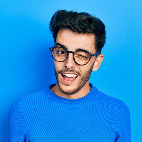 Young man in glasses and blue sweater winking against a solid blue background.