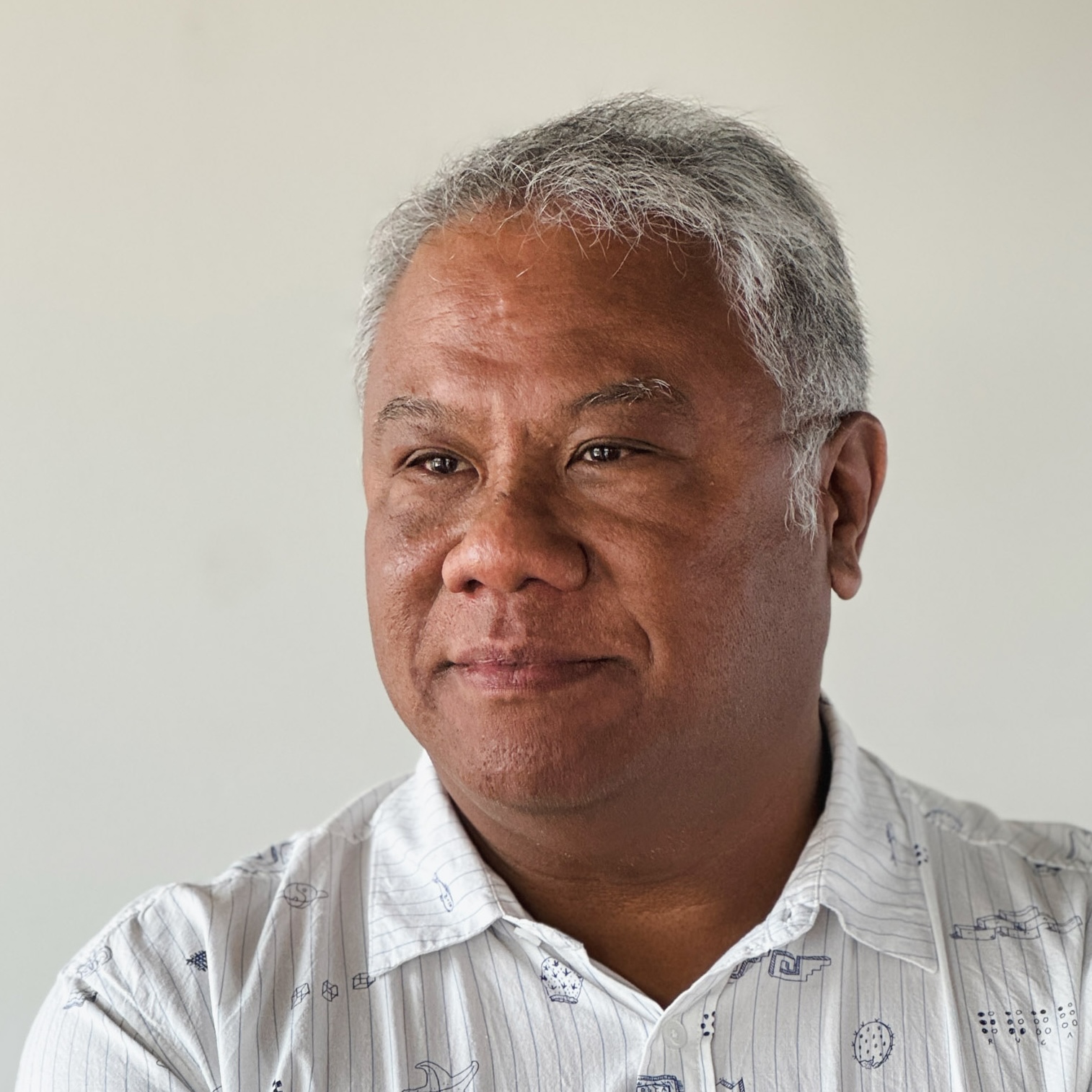 Joe Engalan's headshot. Portrait of a middle-aged man wearing a white patterned shirt smiling slightly against a light background.