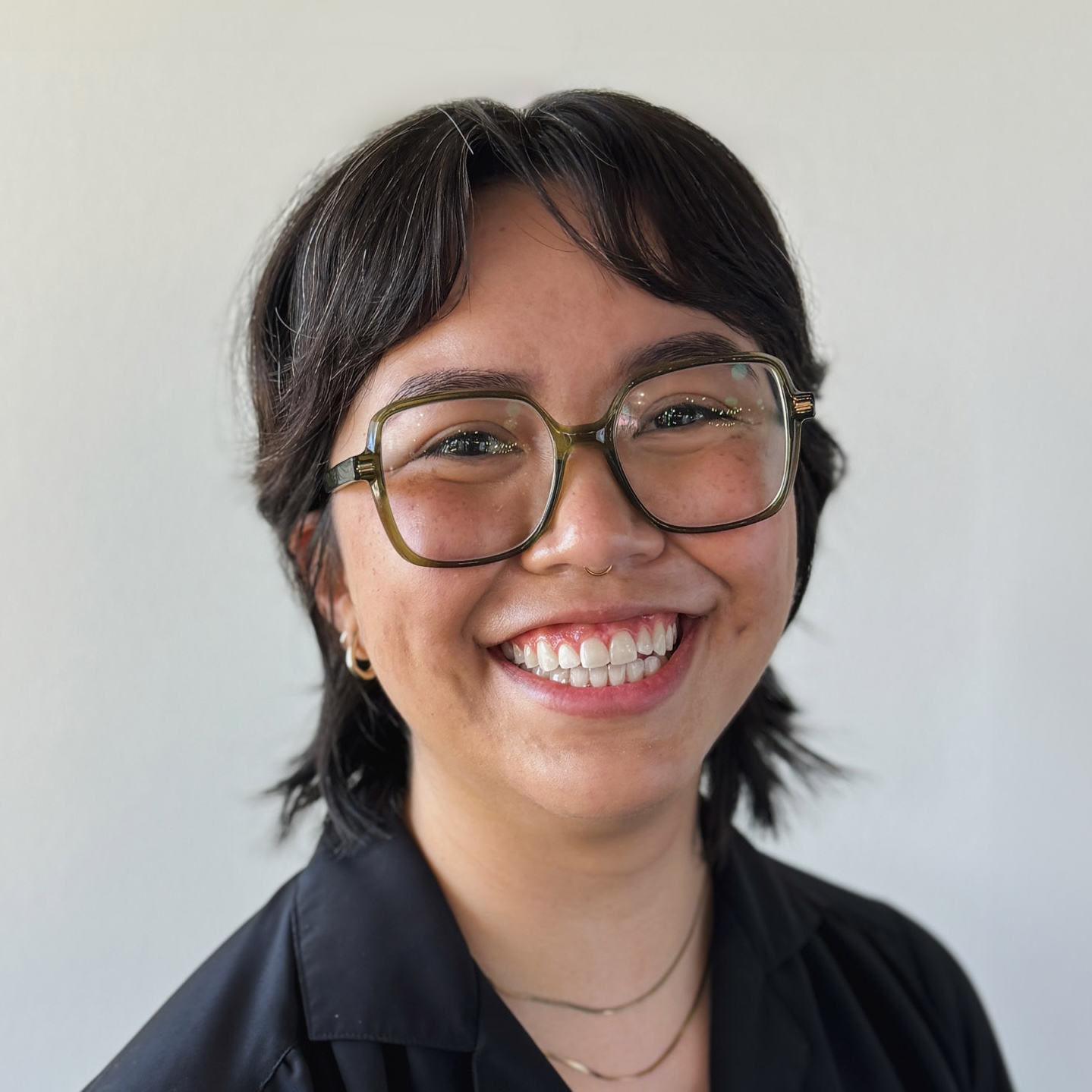Deb Nischik's headshot. Smiling person with short dark hair, large green-framed glasses, gold hoop earrings, and a black shirt.