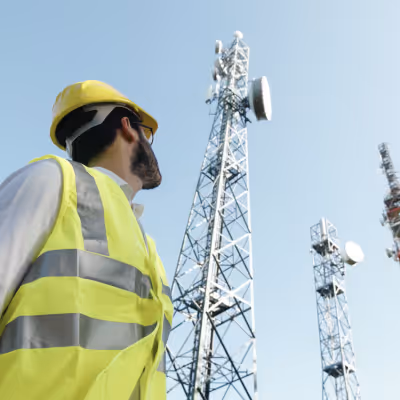 Engineer in safety vest and helmet observing tall communication towers against clear sky.