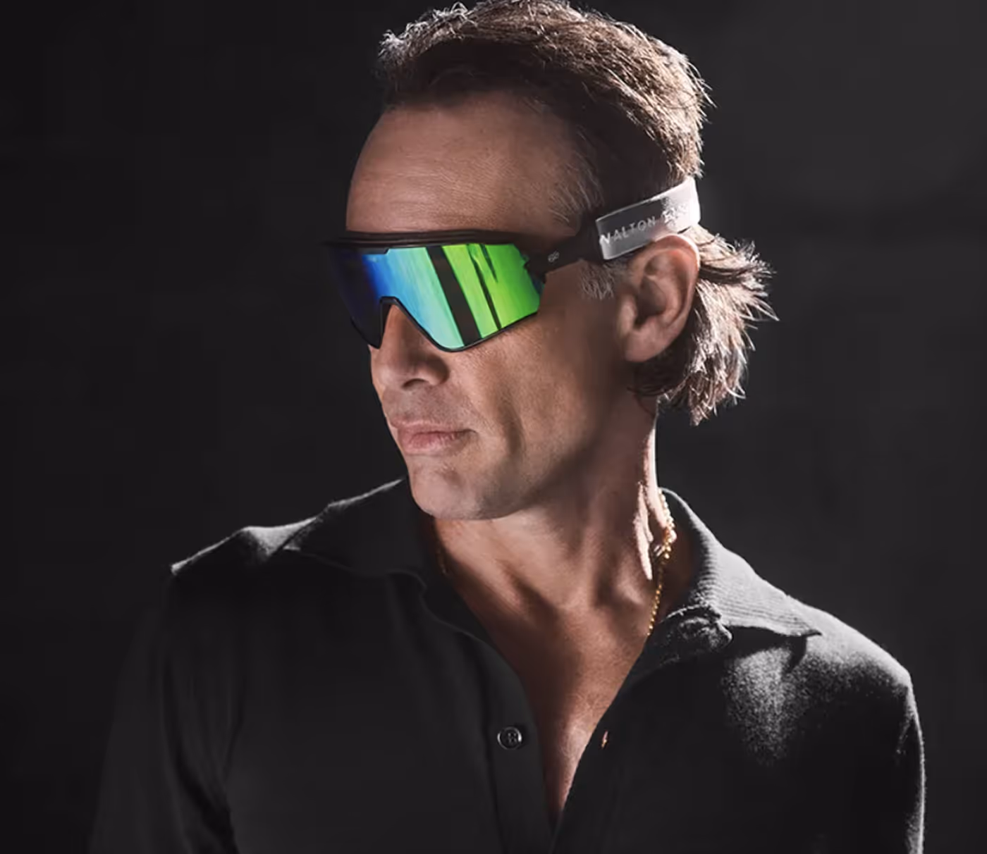 Man wearing black shirt and reflective green sports sunglasses against a dark background.