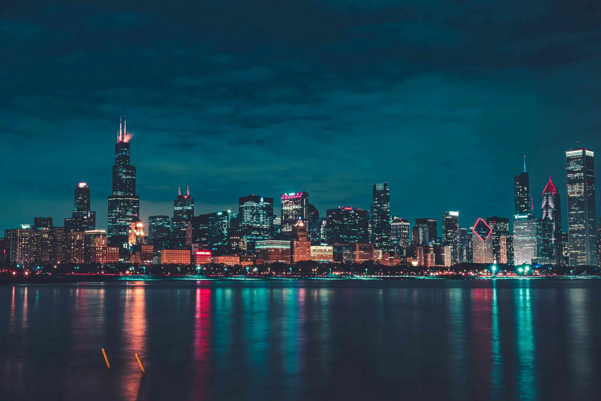 Night cityscape with illuminated skyscrapers reflecting colorful lights on calm water.