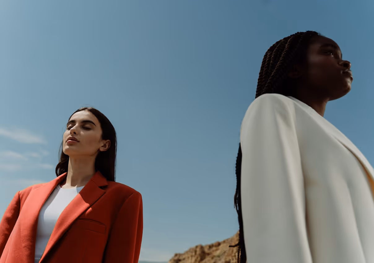 Two women outdoors against a clear sky, one in a red blazer with eyes closed, the other in a white blazer.