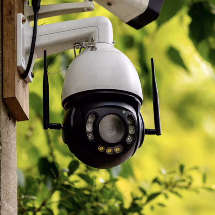 Outdoor white and black security camera mounted on a wooden post with green foliage background.