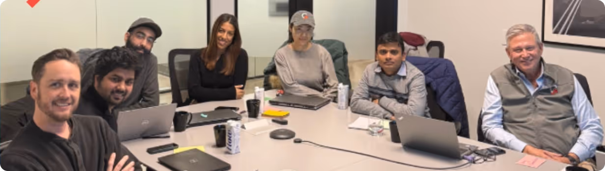 Seven GainShare colleagues sitting around a conference table with laptops and drinks, smiling at the camera.