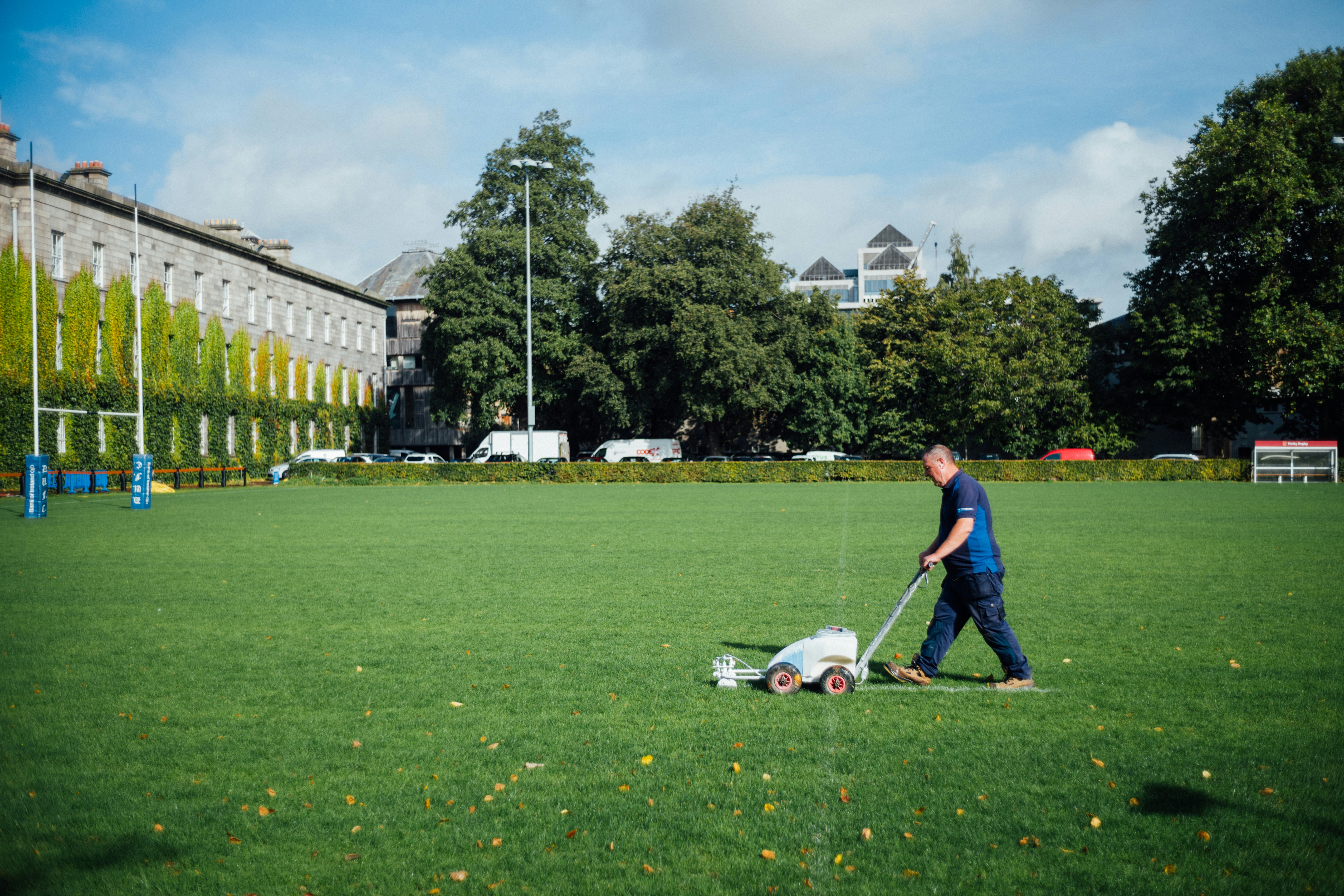 Ein Mann in dunkelblauer Arbeitskleidung schiebt eine weiße Linienmarkierungsmaschine über einen großen, grünen Rasenplatz. Im Hintergrund sind Bäume, ein bewachsenes Steingebäude und Rugby-Pfosten zu sehen
