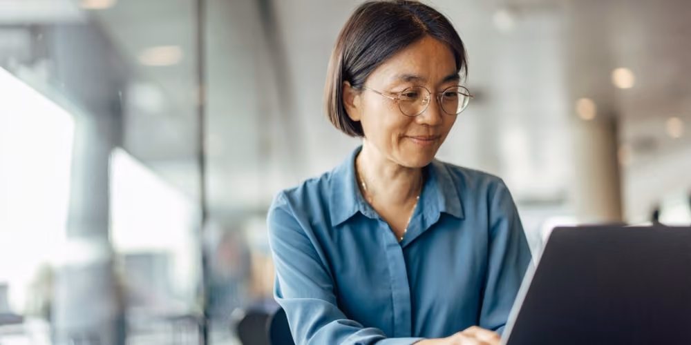 Woman with glasses smiling while working on a laptop in a modern office setting.