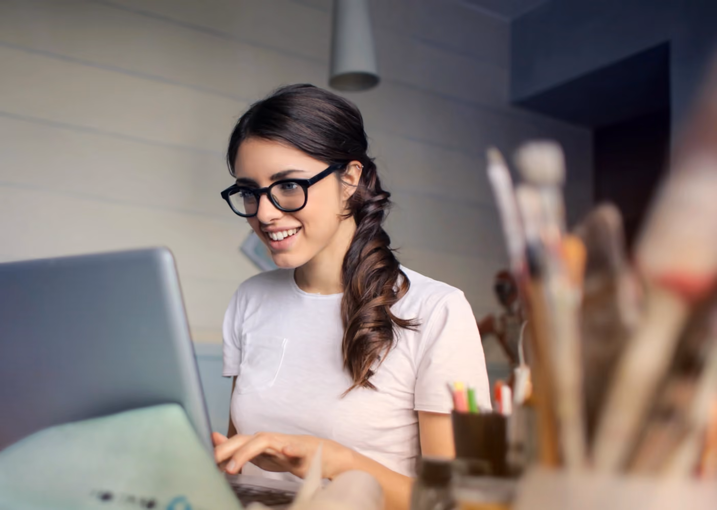 Woman working on a laptop at a home desk, with plants and natural light in the background.