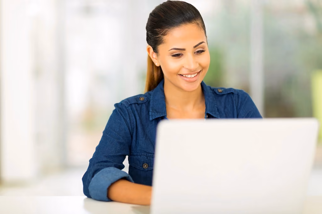 Woman working on a laptop at a home desk, with plants and natural light in the background.