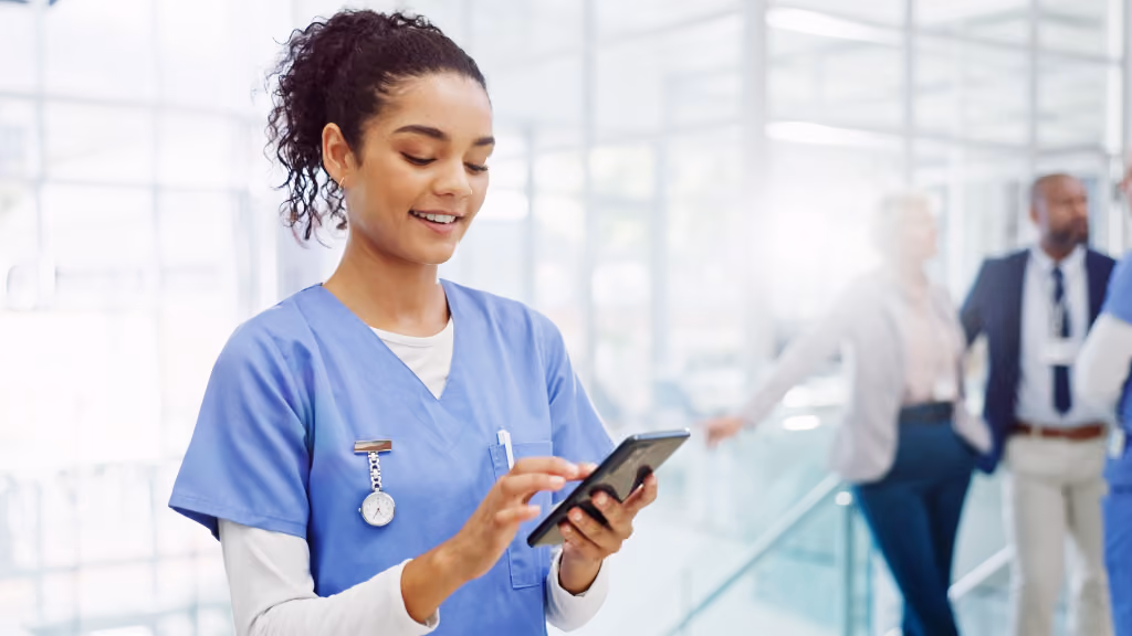 A healthcare professional in scrubs uses a smartphone in a hospital corridor, representing the frontline workers across regulated industries who need mobile-accessible, no-install compliance training that meets them where they are.