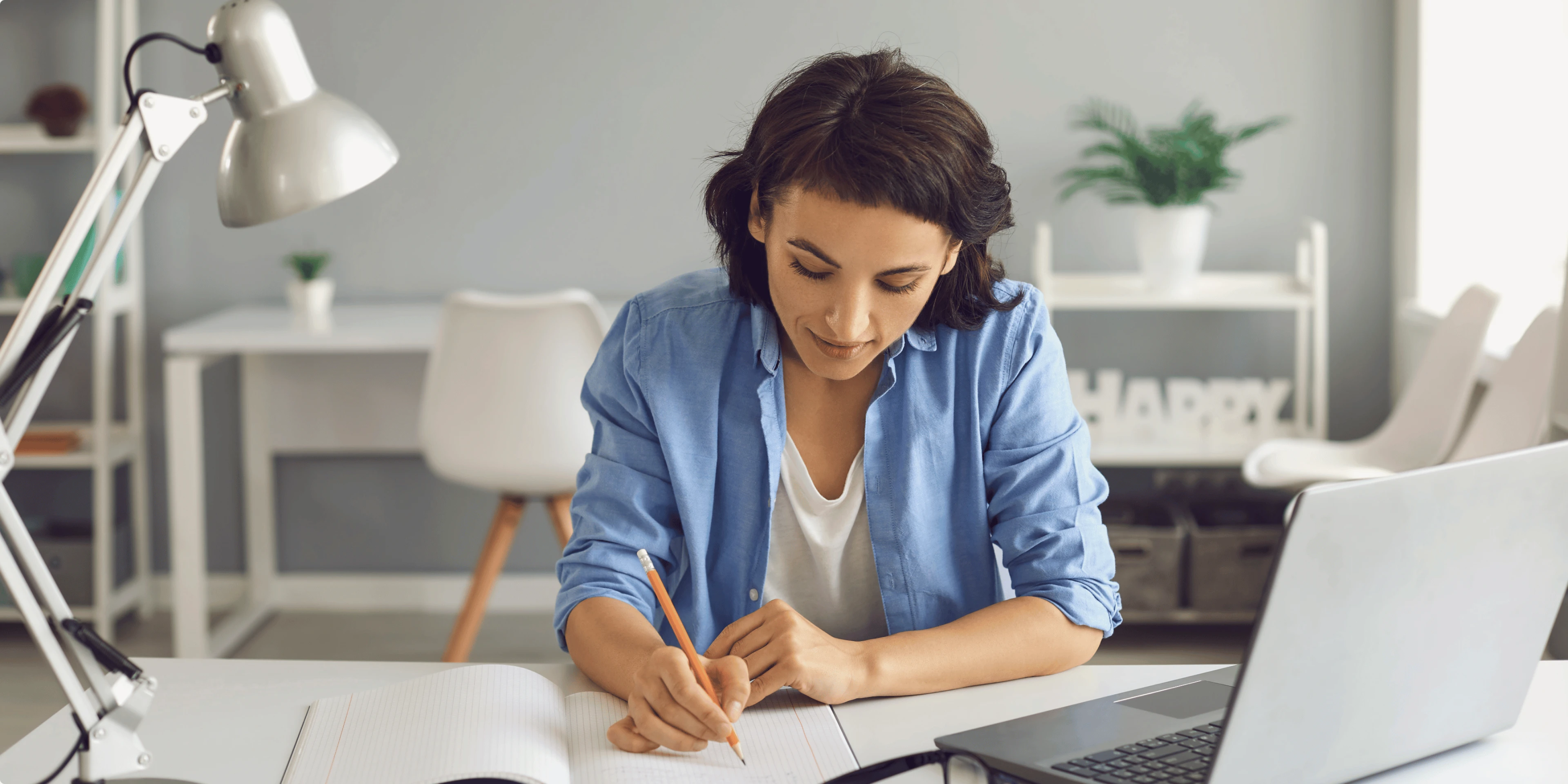 Woman working on a laptop at a home desk, with plants and natural light in the background.