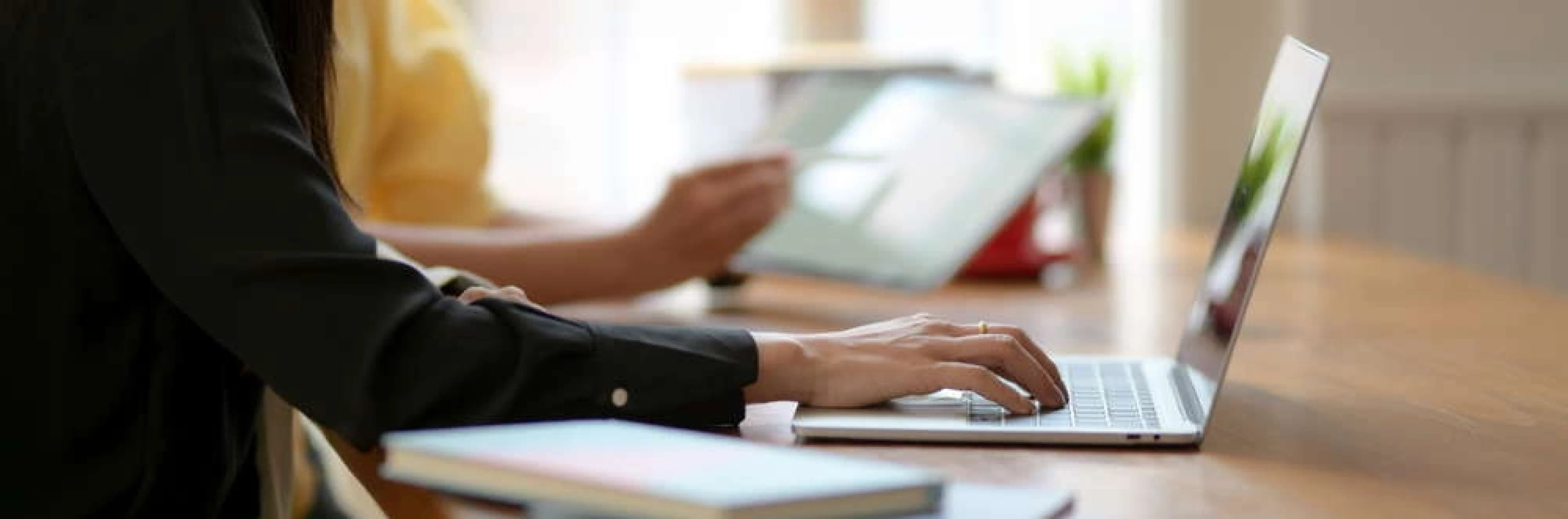 Woman working on a laptop at a home desk, with plants and natural light in the background.
