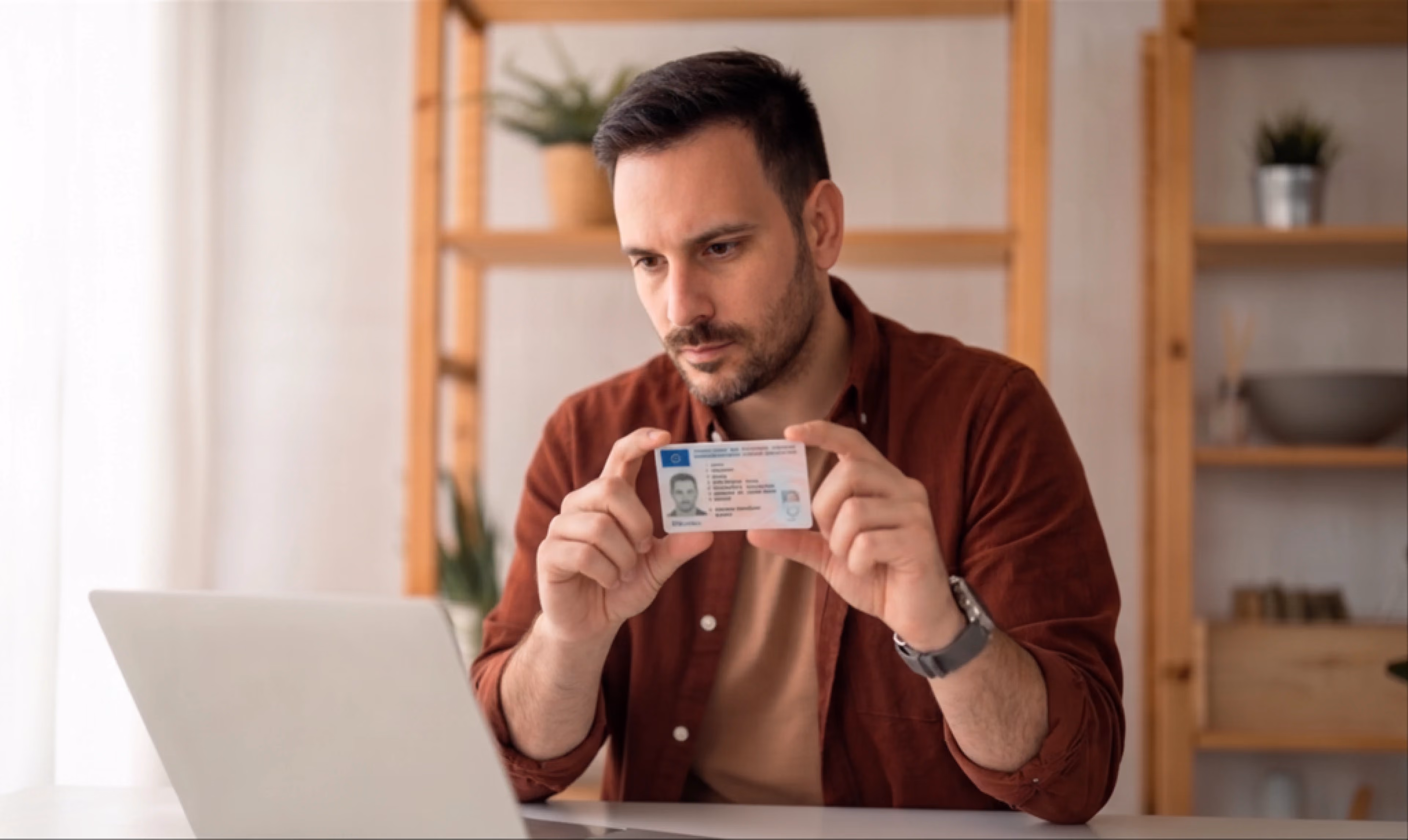 Man holding up an ID card to a laptop camera for identity verification at home.