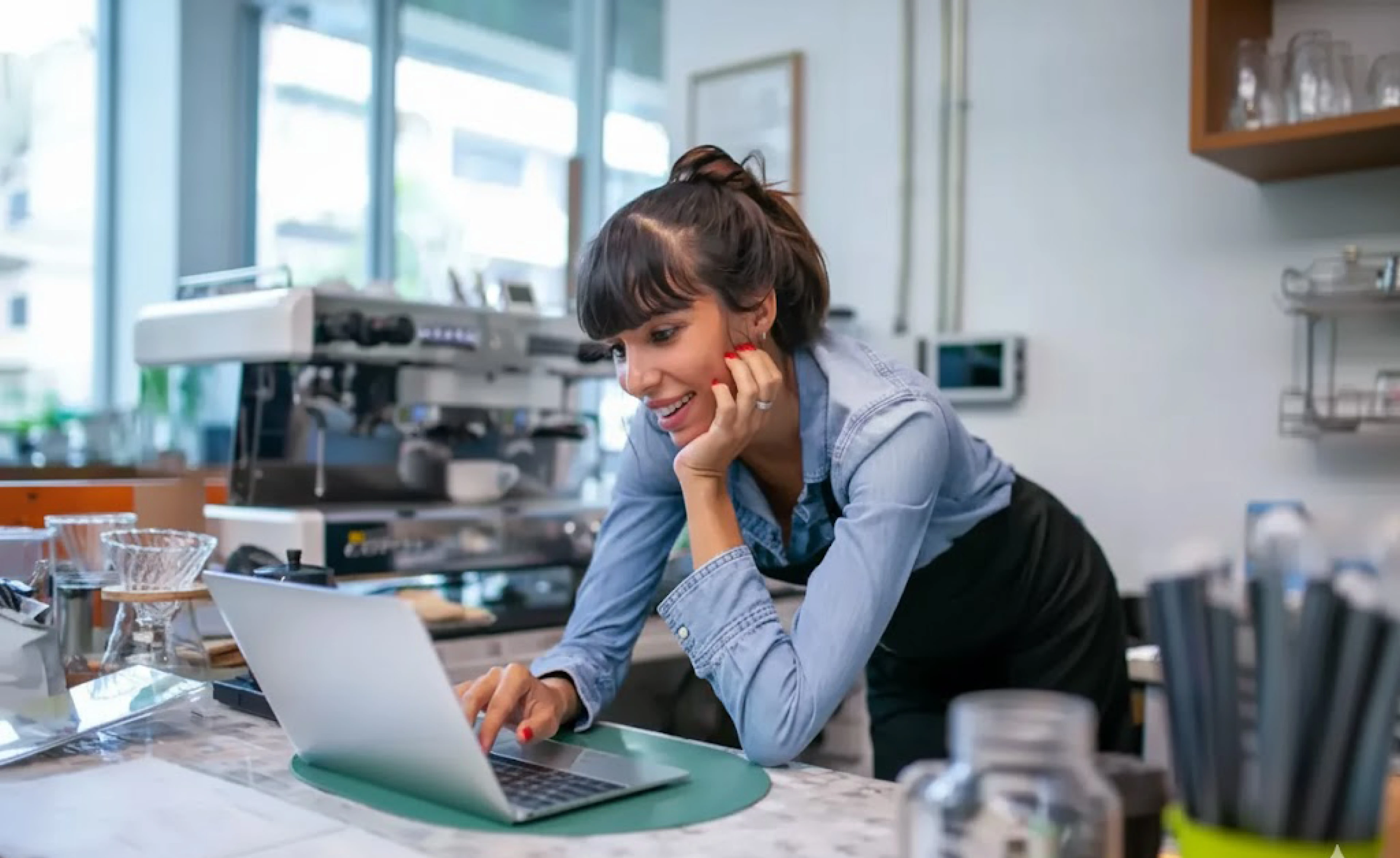 A food service professional in an apron uses a laptop at a cafe counter, representing the hospitality industry workers who complete online food safety and alcohol service certifications through platforms like Integrity Advocate.