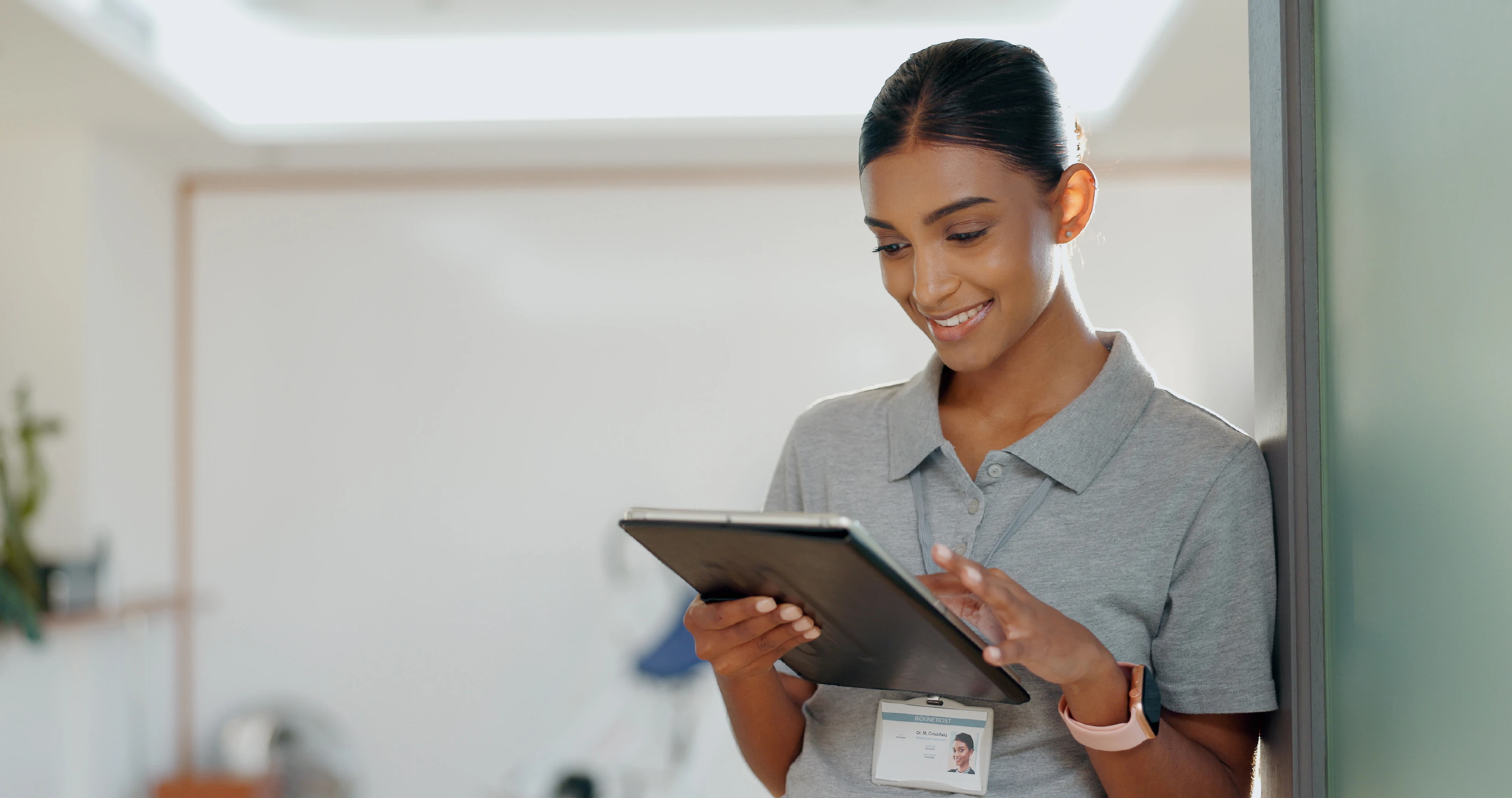 A healthcare professional in scrubs wearing an ID badge uses a tablet in a clinical setting, representing the nurses and clinicians who complete mandatory certification training online and the need for verified, audit-ready credentials in patient care envi