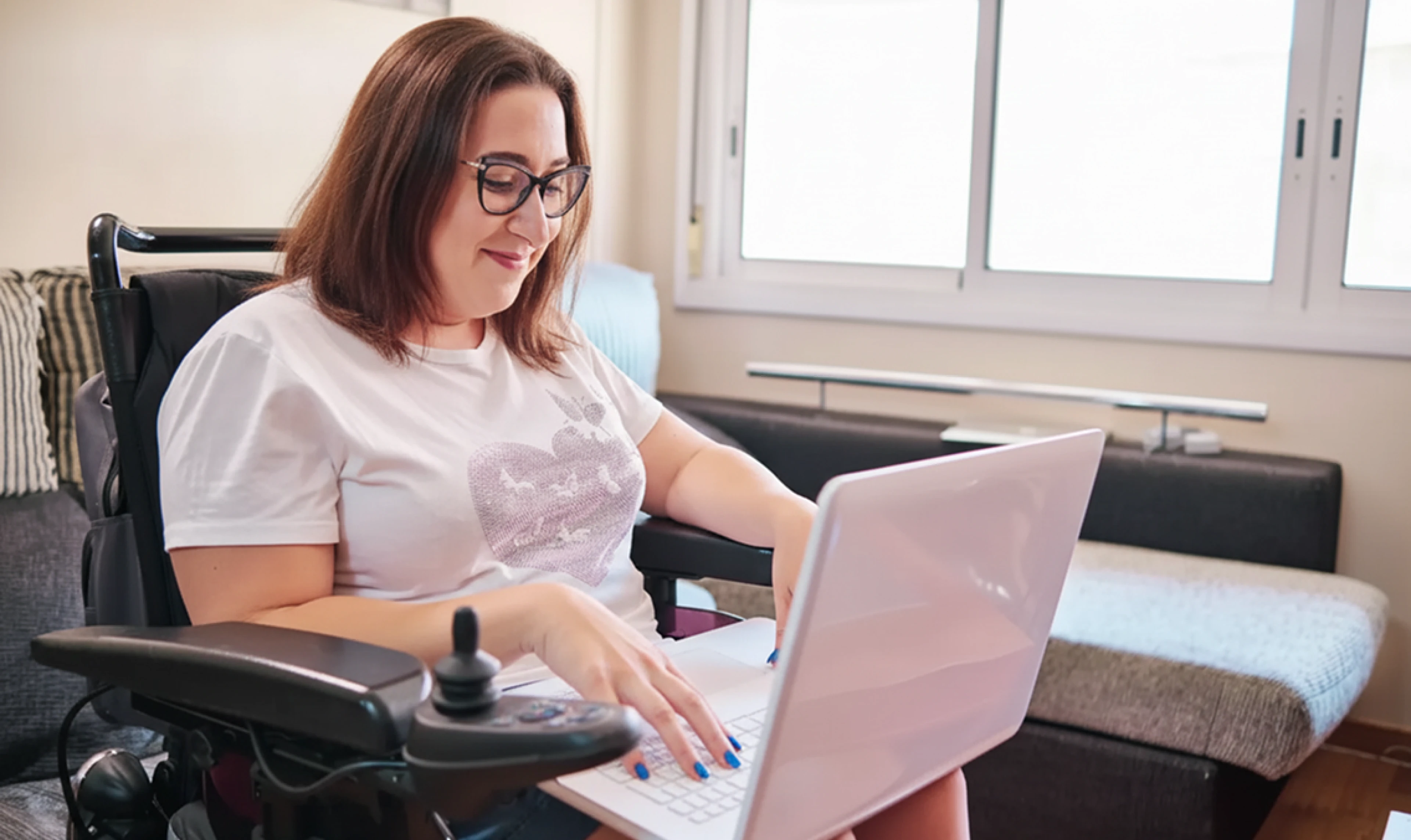 A person using a wheelchair works on a laptop in a comfortable home environment, illustrating accessible online learning and assessment.