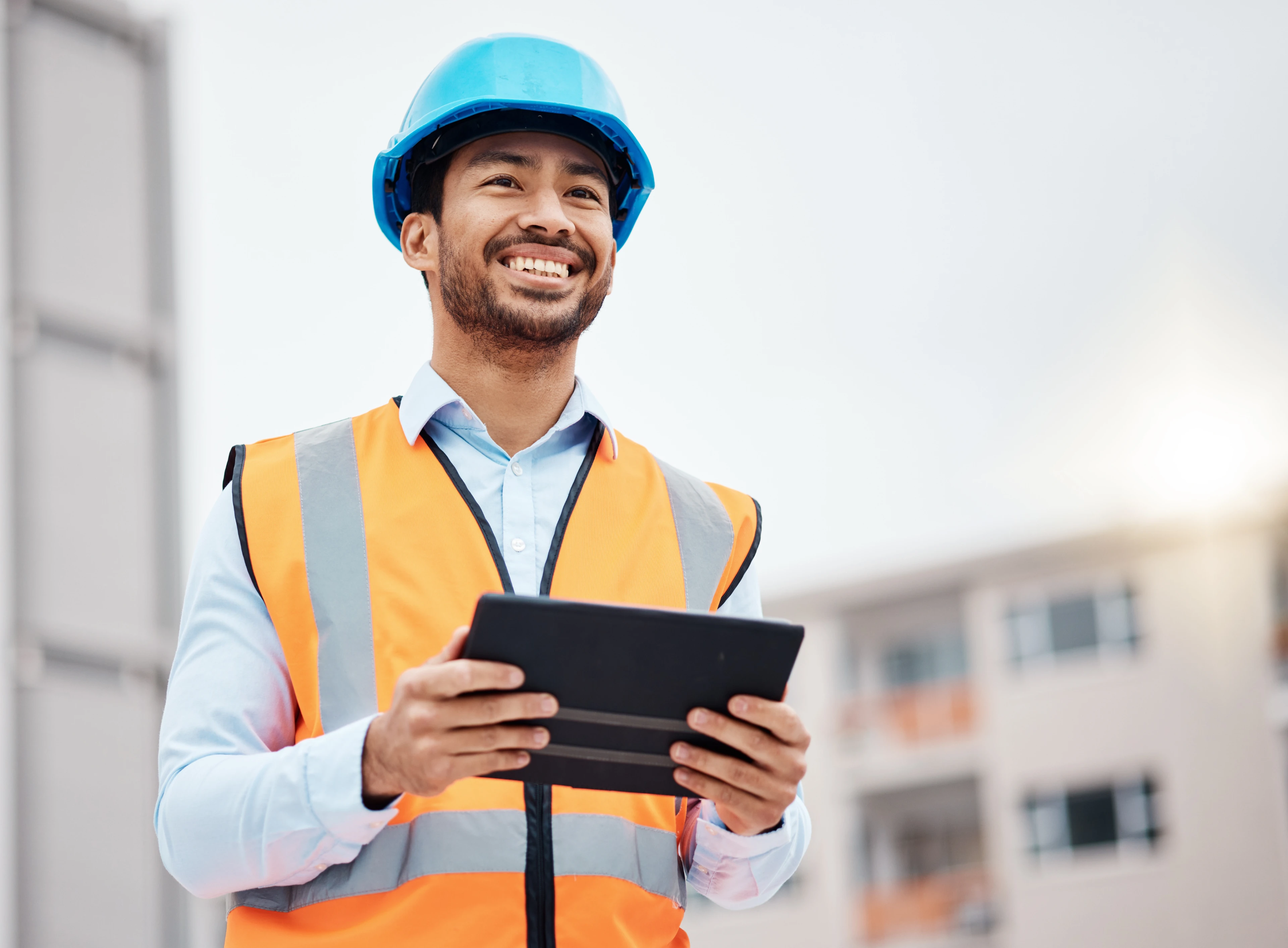 A worker in a hard hat and safety vest reviews a tablet at an industrial job site, representing high-risk workforce training completed through remote proctoring.